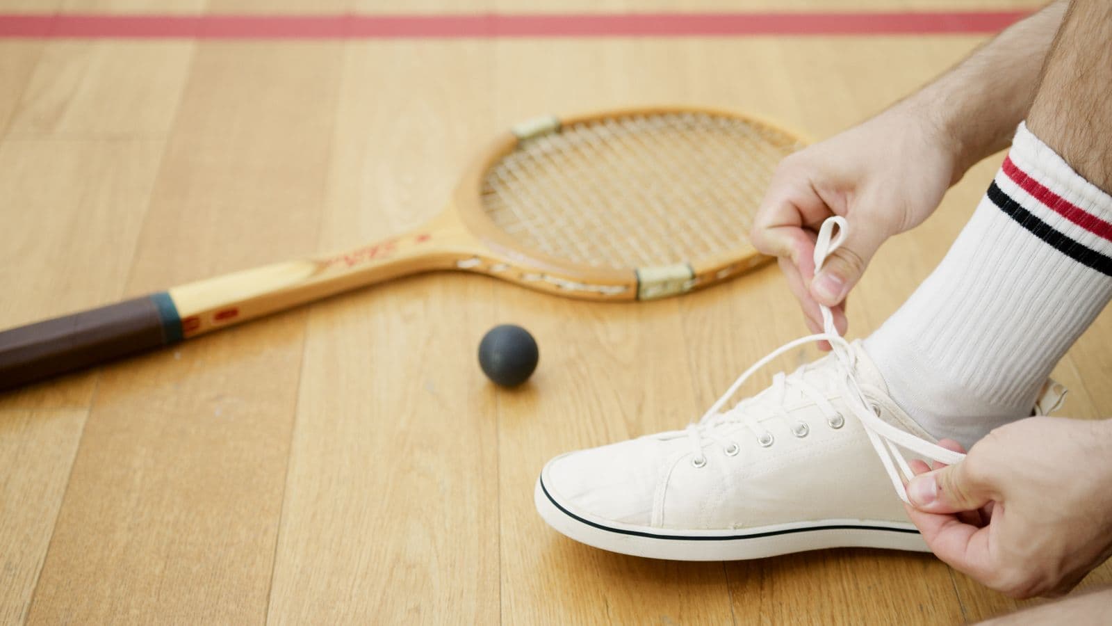 Player tying court shoes on a wooden floor beside a racket
